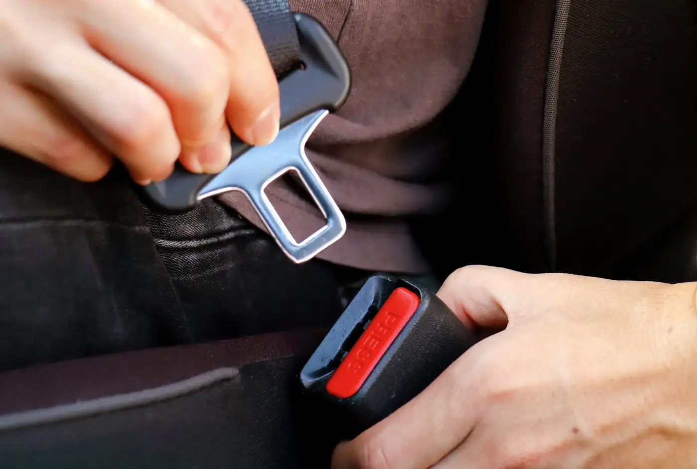 Two hands guiding a seat belt latch tongue into the buckle receptacle inside a vehicle