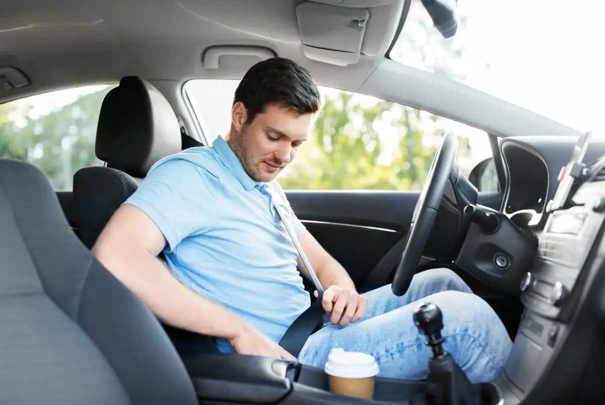 Driver in a blue polo shirt fastening a seat belt in a vehicle cabin with greenery outside the window