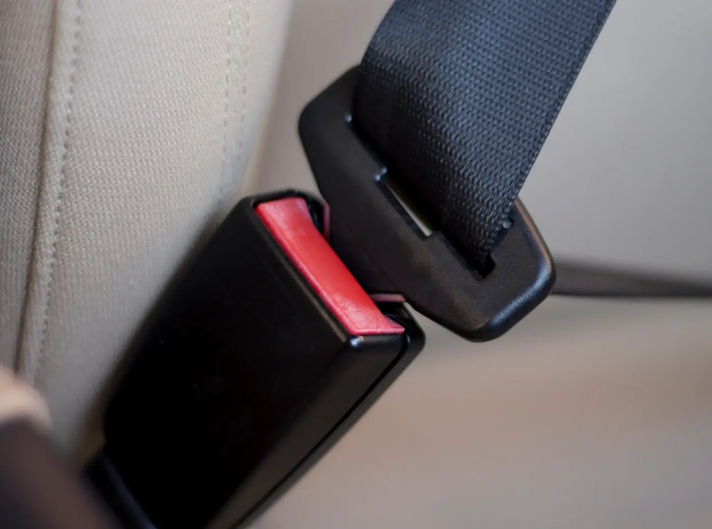 Black seat belt buckle with a red release button beside a latch tongue held in a pillar loop on a beige seat